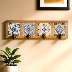 Könnte beinhalten: Ein hölzerner Wandgarderobenständer mit vier dekorativen Keramikfliesen. Jede Fliese zeigt ein einzigartiges blau-weißes Blumenmuster. Der Ständer hat vier bronzefarbene Haken zum Aufhängen von Gegenständen. Der Holzrahmen ist hellbraun.
