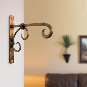 Puede incluir: Soporte de metal de color bronce ornamentado para plantas. El soporte tiene una placa trasera rectangular con dos orificios para tornillos y un diseño de voluta decorativo. El gancho se extiende hacia afuera con un extremo rizado. El acabado tiene una apariencia envejecida y antigua.