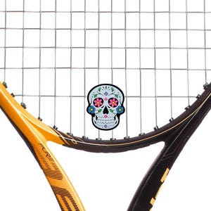 May include: A tennis racket with a black and gold frame and silver strings. A decorative sugar skull sticker with pink, blue, and purple floral accents is centered on the strings. The racket is set against a white background.