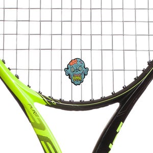 Puede incluir: Una raqueta de tenis con un marco negro y verde neón y un encordado. Una pegatina de cabeza de zombi de dibujos animados está centrada en las cuerdas. La raqueta está en ángulo, mostrando su diseño y el accesorio temático de zombis.