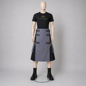Puede incluir: Un maniquí viste una camiseta negra con texto dorado y un kilt gris y negro. El kilt tiene detalles de cadena plateada y botas de cuero negro. El texto de la camiseta dice "Highland Clothes".