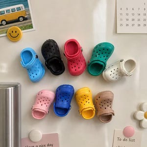 May include: A collection of miniature, colorful clogs in various shades including blue, black, red, green, and white. Additional clogs are pink, blue, yellow, and brown. The clogs are arranged on a white surface, near a calendar and a "To do list" note.