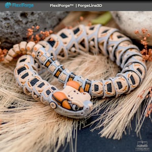 May include: A 3D-printed articulated snake toy in a coiled position. The snake features a segmented design with tan, gray, black, and orange accents. The head has a detailed, robotic appearance. The background includes dried grass and small orange flowers.