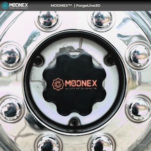 以下が含まれることがあります： 黒いセンターキャップが付いたクロムホイールのクローズアップ。 センターキャップはユニークな形状で、オレンジ色の「MODNEX」ロゴが特徴です。 ホイールには、周囲に複数のクロムラグナットが付いています。