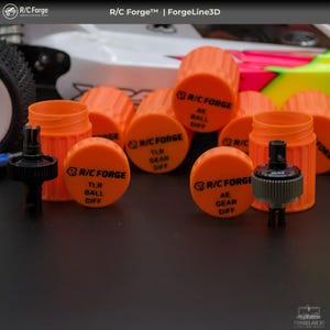 Peut inclure: Plusieurs conteneurs cylindriques orange et des pièces mécaniques noires. Les conteneurs portent l'inscription "R/C Forge" et "AE BALL DIFF". Un objet blanc et vert fluo est visible en arrière-plan.