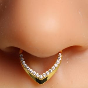 Puede incluir: Un aro de septum dorado en forma de V, adornado con pequeñas piedras preciosas transparentes. La joya se muestra en la nariz de una persona, destacando el diseño y el brillo. Este aro de septum es un tipo de joya corporal.