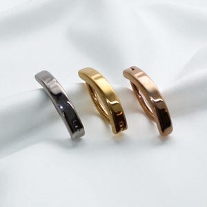 May include: Three metal hair clips in silver, gold, and rose gold. The clips are curved and have a simple, elegant design. They are arranged on a white, textured surface, creating a minimalist aesthetic. The clips are likely for styling or securing hair.