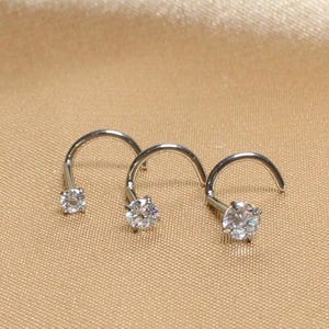 May include: Three silver nose studs with clear, round gemstones. The studs are in a curved screw shape, with the gemstone at the end of the screw. The background is a light tan color.