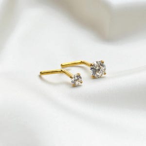 May include: A pair of gold-colored stud earrings with clear, round gemstones. The earrings are set on a white, silky background, creating a contrast that highlights the jewelry's sparkle and design. The earrings are angled, showing their post and stone settings.