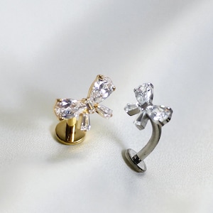 May include: Two bow-shaped stud earrings. One is gold-toned with clear, sparkling stones. The other is silver-toned with similar clear stones. Both earrings are on a white background.