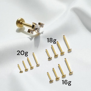 May include: Gold-colored body jewelry, including a labret stud with a clear gem cluster and several straight barbell studs in 16g, 18g, and 20g sizes. The jewelry is displayed on a white, textured surface.