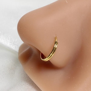 May include: A close-up of a gold nose ring. The ring is a small hoop with a double-band design. The jewelry is worn on a person's nose. The skin tone is light. The background is a white fabric.