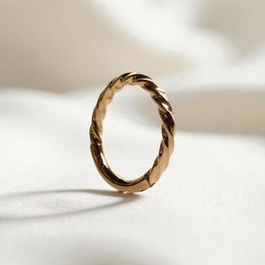 Puede incluir: Un anillo dorado con un diseño de cuerda trenzada. El anillo tiene forma ovalada y está sobre una superficie blanca texturizada. El diseño del anillo es detallado y refleja la luz.