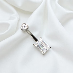 Puede incluir: Un piercing de ombligo plateado con una gema rectangular transparente grande en la parte inferior y una gema redonda más pequeña en la parte superior. La joya está sobre un fondo blanco y sedoso.