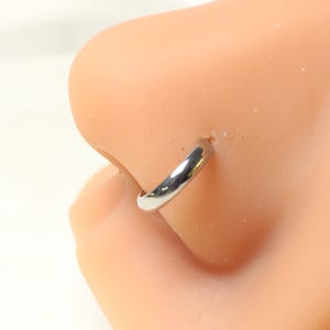 May include: A silver nose ring is shown in a close-up shot. The ring is a simple, curved band, and it is inserted into a nose piercing. The skin around the nose is a light peach color.