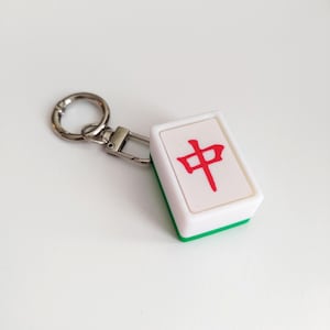 Op de afbeelding: Een witte en groene Mahjong-tegel sleutelhanger met een zilveren sluiting en ring. De tegel heeft een rood Chinees karakter. Een leuk accessoire voor spelliefhebbers.