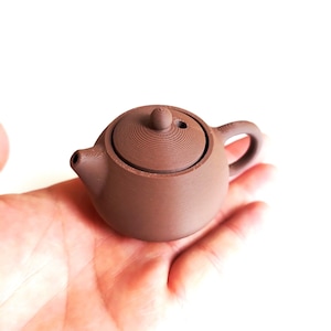 May include: A miniature brown teapot with a lid, spout, and handle. The teapot is held in a hand against a white backdrop. The teapot has a rounded body and a small, circular lid with a knob.