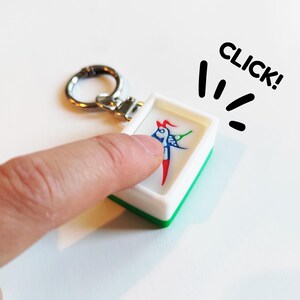 Pu&ograve; includere: Un portachiavi bianco e verde con un'illustrazione colorata di una figura simile a un uccello. Il portachiavi viene premuto con un dito. La parola "CLICK!" &egrave; scritta in nero.