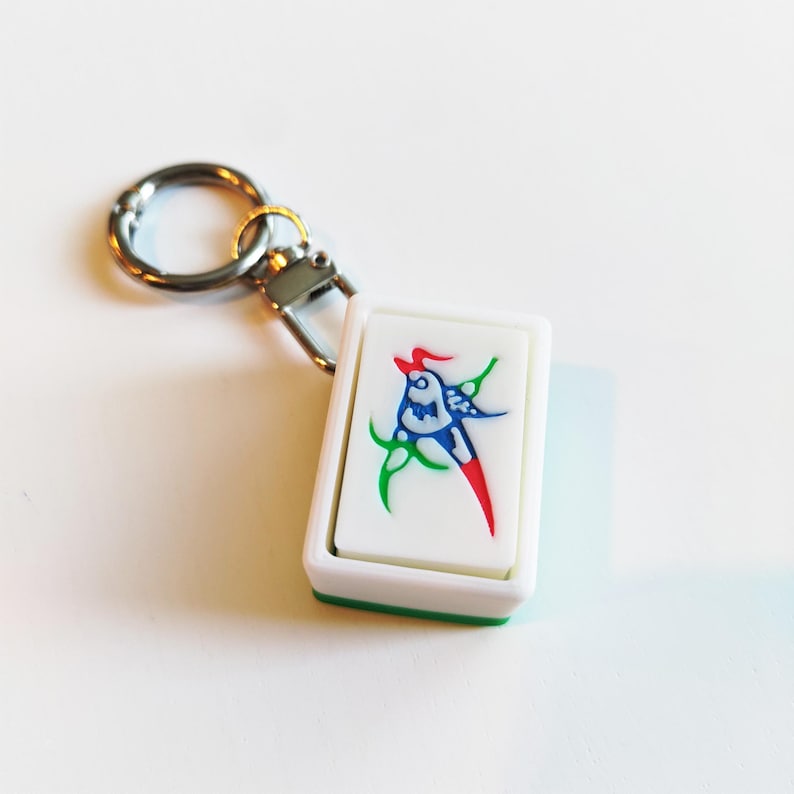 Pu&ograve; includere: Un portachiavi rettangolare bianco con un design colorato con accenti rossi, blu e verdi. Il portachiavi ha un anello e un fermaglio in metallo argentato. Il design sembra essere un uccello stilizzato o una creatura mitica.