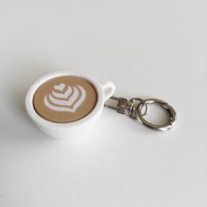 Könnte beinhalten: Ein weißer Kaffeebecher-Schlüsselanhänger mit Latte-Art-Design. Der Schlüsselanhänger zeigt ein detailliertes, hellbraunes Kaffee-Design mit einem weißen herzförmigen Muster. Er hat einen silbernen Verschluss und Ring.