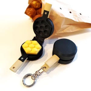 May include: A black and cream waffle iron keyring with a yellow waffle. The keyring is next to a closed version of the waffle iron and a paper bag with round pastries.