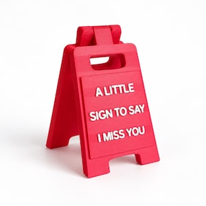 May include: A bright red A-frame sign featuring a cutout handle at the top. The front panel displays white raised lettering that reads A LITTLE SIGN TO SAY I MISS YOU. The structure is freestanding with a hinged top and a matte finish.