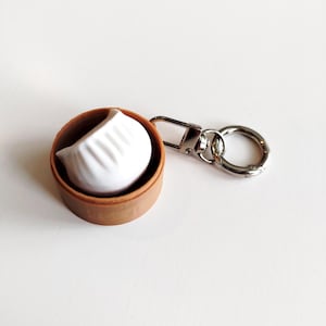 May include: A white dumpling keyring inside a small, round, brown bamboo steamer. The dumpling has detailed pleats. The keyring has a silver clasp and a silver ring.