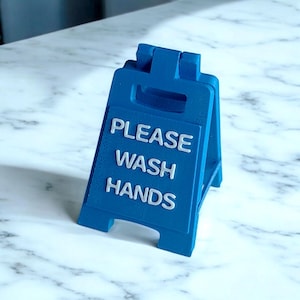 Peut inclure: Un panneau bleu en forme de A avec les mots "PLEASE WASH HANDS" en lettres blanches. Le panneau est fait d'un matériau solide et est conçu pour tenir debout. Le panneau est sur une surface en marbre.