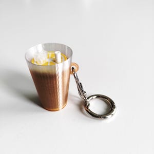 May include: A novelty keychain shaped like an iced beverage. The cup is a gradient of brown to clear, with a yellow lemon slice and ice cubes visible inside. A silver-colored clasp and ring are attached.