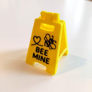 May include: A small, bright yellow A-frame sign featuring the text "BEE MINE" in black, accompanied by a heart and a bee illustration. The plastic sign is designed to stand on its own, suitable for display.