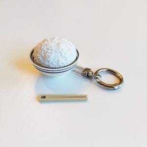 May include: A novelty keychain with a miniature bowl of white rice, blue stripes, a silver-tone clasp, and a small, beige ruler. The rice has a textured appearance. The items are on a white surface.