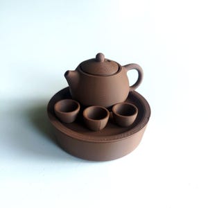 May include: A brown ceramic tea set featuring a teapot with a lid, three small cups, and a round tray. The teapot has a curved handle and spout. The set is designed for serving tea.
