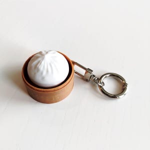 May include: A white ceramic dumpling keychain in a brown bamboo steamer. The keychain has a silver-coloured clasp and a round key ring. The dumpling has detailed pleats, and the steamer has a textured appearance.