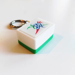 Pu&ograve; includere: Un portachiavi con tessera Mahjong bianca e verde con un portachiavi argentato. La tessera presenta un design colorato con accenti rossi, verdi e blu. Il portachiavi &egrave; di forma rettangolare.
