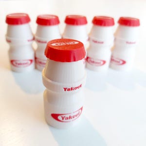 May include: A group of small, white Yakool yogurt drink bottles with red lids. The bottles are arranged together, with the Yakool logo in red on the front. The image is taken in bright light.