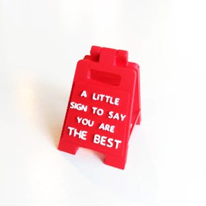 May include: A bright red, miniature sign with the text "A LITTLE SIGN TO SAY YOU ARE THE BEST" in white. The sign is shaped like a small, folding A-frame, perfect for a desk or shelf.