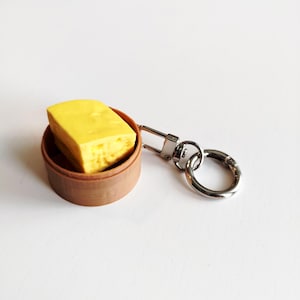 May include: A novelty keychain with a miniature dim sum item. The yellow, rectangular food is in a small brown container. The keychain has a silver clasp and ring, ideal for keys or bags.