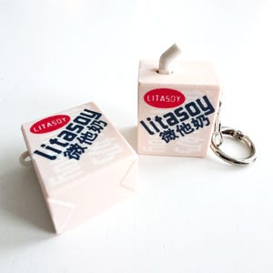 Puede incluir: Dos llaveros en miniatura de cartón de leche de soja Litasoy de color rosa claro. Cada cartón presenta el nombre de la marca en inglés y chino, con un logotipo ovalado rojo. Un llavero tiene un anillo plateado.