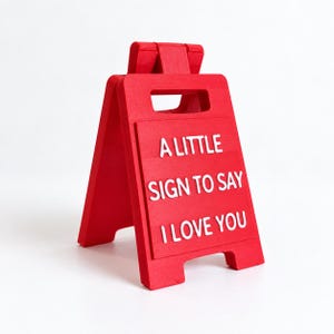 May include: A bright red A-frame sign featuring a cutout handle at the top. The front panel displays raised white lettering that reads A LITTLE SIGN TO SAY I LOVE YOU. The structure is hinged at the top, allowing it to stand independently.