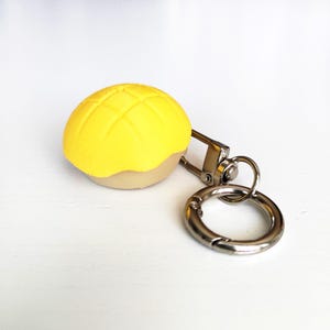 Pode incluir: Chaveiro de pão de melão, um pão doce japonês. A parte superior é amarela com um padrão quadriculado, e a base é bege. Possui um fecho e um anel prateados.