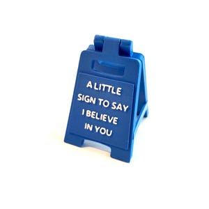 May include: A blue plastic sign with white text reading "A LITTLE SIGN TO SAY I BELIEVE IN YOU". The sign is shaped like an A-frame. The background is plain white.