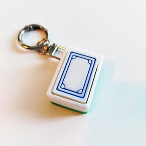 Op de afbeelding: Een witte en blauwe rechthoekige sleutelhanger met een zilveren ring. De sleutelhanger heeft een blauw omrand ontwerp op een witte achtergrond, met een groene rand. De ring is bevestigd aan een metalen sluiting.