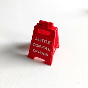 Puede incluir: Un pequeño letrero rojo brillante en forma de A con el texto blanco "A LITTLE SIGN FULL OF HUGS". El letrero es de plástico y está sobre una superficie blanca.