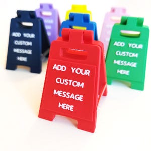 Puede incluir: Una colección de coloridos letreros en forma de A en rojo, verde, azul, morado y amarillo. Cada letrero tiene el texto blanco "ADD YOUR CUSTOM MESSAGE HERE", perfecto para mensajes personalizados.