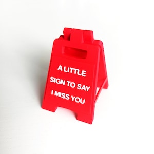 May include: A bright red, plastic, folding sign with the text "A LITTLE SIGN TO SAY I MISS YOU" in white. The sign is designed to stand upright, perfect for a desk or shelf. The sign is a simple, yet heartfelt, message.