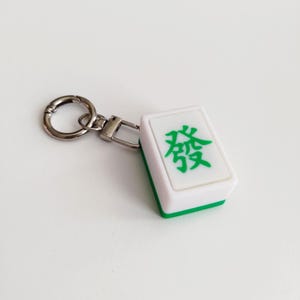 May include: A white and green Mahjong tile keychain with a silver-colored clasp and key ring. The tile features a green Chinese character. The keychain is a fun accessory for game enthusiasts.