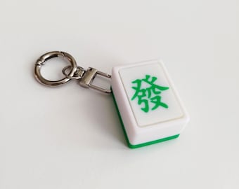 May include: A white and green Mahjong tile keychain with a silver-colored clasp and key ring. The tile features a green Chinese character. The keychain is a fun accessory for game enthusiasts.