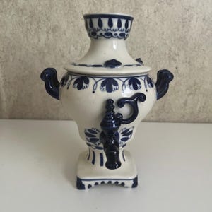 May include: A ceramic samovar featuring a white base with intricate dark blue floral patterns. The vessel includes two side handles, a central spigot, a removable lid, and a square pedestal base, all decorated with matching blue painted motifs.