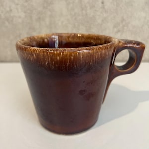 May include: A brown ceramic mug with a unique handle design. The mug has a dark brown exterior with a lighter brown rim. The handle features a circular opening. The mug is likely vintage or retro.