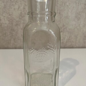 May include: Clear glass honey bottle with a rectangular body and a cylindrical neck. The bottle features an embossed image of a beehive and the words "1 POUND PURE HONEY". The bottle is empty and sits on a white surface.
